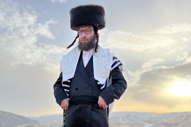 Rabbi Akiva Weingarten steht in der Wüste in der Nähe Jerusalems auf einem Felsen und ist in traditionellen, ultraorthodoxen Gewändern gekleidet. Er trägt einen dunkelbraunen Schtreimel (Kopfbedeckung), einen schwarzen Kaftan und einen weiß-schwarzen Tallit (Gebetsmantel). Er trägt Vollbart und Schläfenlocken. Im Hintergrund ist die untergehende Sonne und eine karge Wüstenlandschaft zu erkennen.
