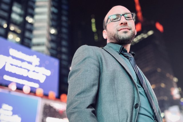 m Vordergrund des Bildes ist ein modern gekleideter Rabbi Akiva in einem türkisenen Anzug zu sehen. Er steht nachts auf dem Time Square in New York; im Hintergrund sind Menschen und Skyscraper in der Unschärfe zu erkennen.