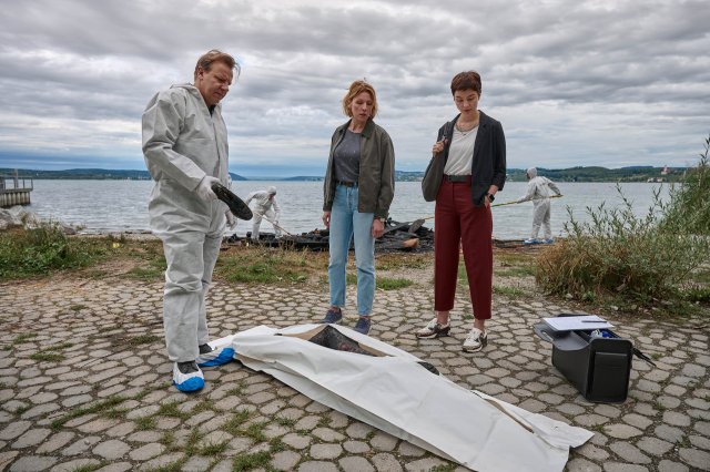 "Mordufer - Brennen soll sie": Gregor Eisen (Ronald Kukulies), Doro Beitinger (Franziska Weisz) und Chiara Locatelli (Maria Wördemann) stehen am Ufer des Bodensees an einem Tatort. Vor ihnen auf dem Boden liegt eine Brandleiche in einem Leichensack. Im Hintergrund sind Spurensicherer bei der Arbeit und suchen das Ufer ab. Gregor Eisen und die Spurensicherer tragen weiße Overalls, blau-weiße Überzieher über ihren Schuhen und weiße Handschuhe.