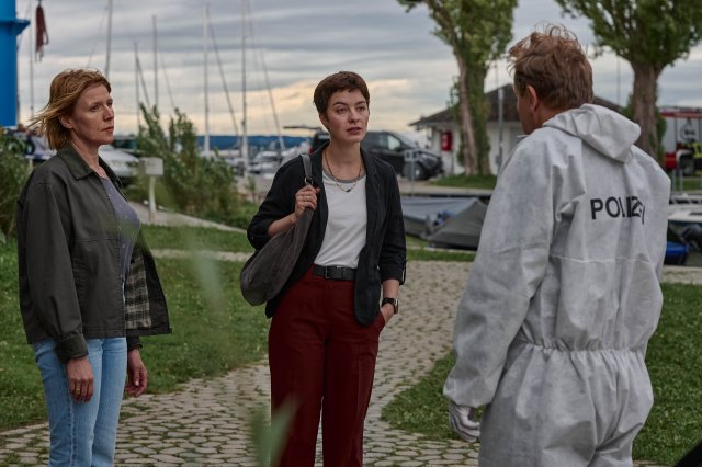 "Mordufer - Brennen soll sie": Doro Beitinger (Franziska Weisz), Chiara Locatelli (Maria Wördemann) und Gregor Eisen (Ronald Kukulies) stehen auf einer Grünfläche am Rande des Bodensees und unterhalten sich. Gregor Eisen trägt einen weißen Overall und weiße Handschuhe. Im Hintergrund liegen einige Boote im Wasser.
