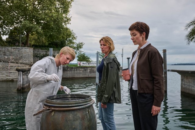 "Mordufer - Kurzes Glück": Sarah Hertweg (Anna Gesa-Raija Lappe), Doro Beitinger (Franziska Weisz) und Chiara Locatelli (Maria Wördemann) stehen am Ufer des Bodensees vor einem offenen Fass. Hertweg trägt einen weißen Overall und weiße Handschuhe, mit der rechten Hand hält sie eine Spritze über das Fass. Beitinger und Locatelli beobachten sie dabei.