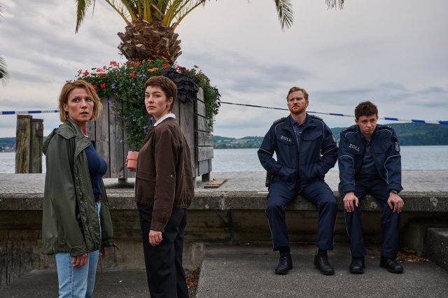 "Mordufer - Kurzes Glück": Doro Beitinger (Franziska Weisz) und Chiara Locatelli (Maria Wördemann) stehen auf der Uferpromenade am Rande des Bodensees. Rechts hinter ihnen sitzen Florian Jäger (Lucas Prisor) und Nico Arnold (Paul Ahrens) auf einer Mauer, beide tragen Zoll-Uniformen. Alle vier Personen blicken mit entsetzten Mienen nach vorne.