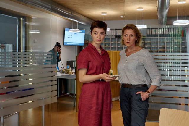 "Mordufer - Die große Gier": Chiara Locatelli (Maria Wördemann) und Doro Beitinger (Franziska Weisz) stehen in einem Büro und blicken zur Seite. Locatelli hält ein Smartphone in ihrer rechten Hand. Im Hintergrund steht Katrin Glaunsinger (Beate Constanze Pitronik) an einem Schreibtisch und telefoniert mit einem Festnetztelefon.