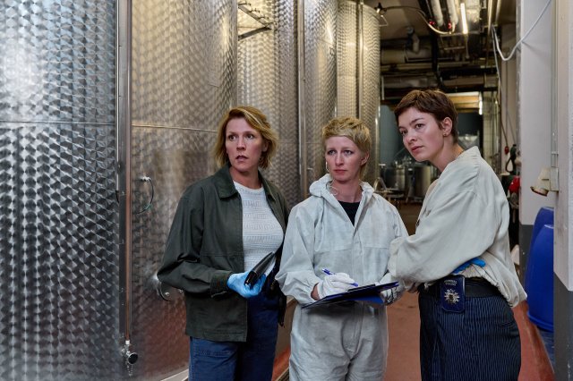 "Mordufer - Wein und Wahrheit": Doro Beitinger (Franziska Weisz), Sarah Hertweg (Anna Gesa-Raija Lappe) und Chiara Locatelli (Maria Wördemann) stehen im Keller eines Weinguts vor großen silbernen Tanks und blicken interessiert zur Seite. Sie tragen Handschuhe, Beitinger hält eine Geldbörse in ihrer rechten Hand, Hertweg hält ein Klemmbrett in ihrer linken Hand und einen Stift in ihrer rechten Hand. Hertweg trägt einen weißen Overall der Spurensicherung.