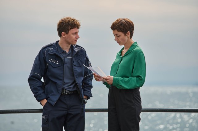 "Mordufer - Wein und Wahrheit": Nico Arnold (Paul Ahrens) steht mit Chiara Locatelli (Maria Wördemann) am Ufer des Bodensees vor einem Geländer. Er blickt sie erwartungsvoll an, während sie auf ein Schriftstück schaut, welches sie in ihren Händen hält.