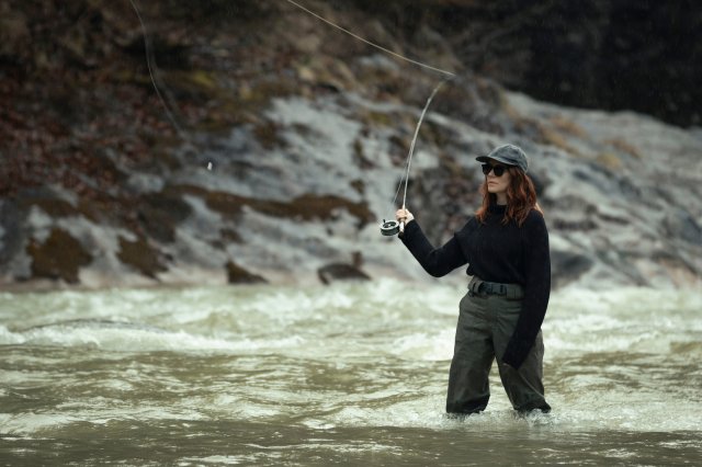 “Der Garmisch-Krimi - Wolfsmord“: Ira Zach (Lavinia Wilson) steht in einem knietiefen Gewässer. Sie trägt eine Gummihose, einen schwarzen Pullover, ihre schwarze Sonnenbrille und eine Kappe auf dem Kopf. In der Hand hält sie ihre Angel, mit der sie gerade zum Wurf ausholt.