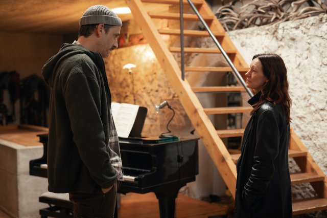 “Der Garmisch-Krimi - Wolfsmord“: Ira Zach (Lavinia Wilson) und ihr Expartner Johann Simader (Max von Thun) stehen sich in einem Wohnzimmer vor einer Steinwand gegenüber und schauen sich tief in die Augen. Im Hintergrund steht ein schwarzer Flügel vor einer Holztreppe.