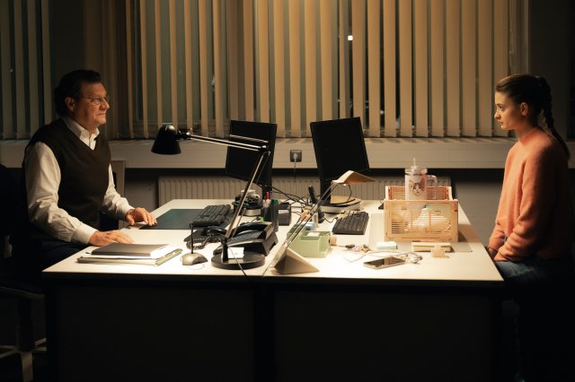 “Der Garmisch-Krimi - Wolfsmord“: Daphne Meindl (Philine Schmölzer) und Kommissariats-Chef Graner (Harald Windisch) sitzen sich an einem Schreibtisch auf dem Kommissariat gegenüber. Draußen ist es bereits dunkel geworden und die Jalousien zugezogen. Vor Daphne Meindl steht eine Kiste, in die sie für den Fall einer Suspendierung bereits ihren fliederfarbenen Trinkbecher, sowie ihre Schreibtischdeko gepackt hat.